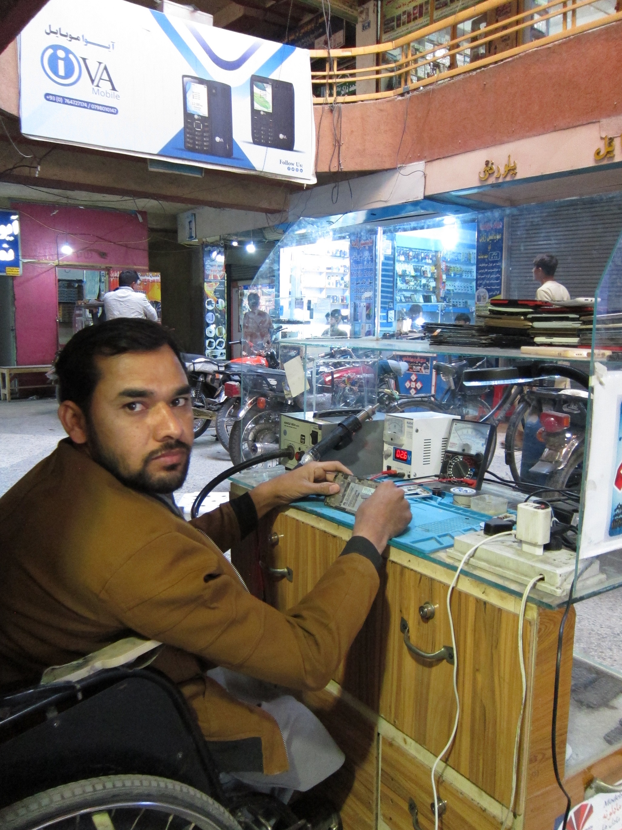 A man in a wheelchair looks at the camera as he repairs a mobile phone.