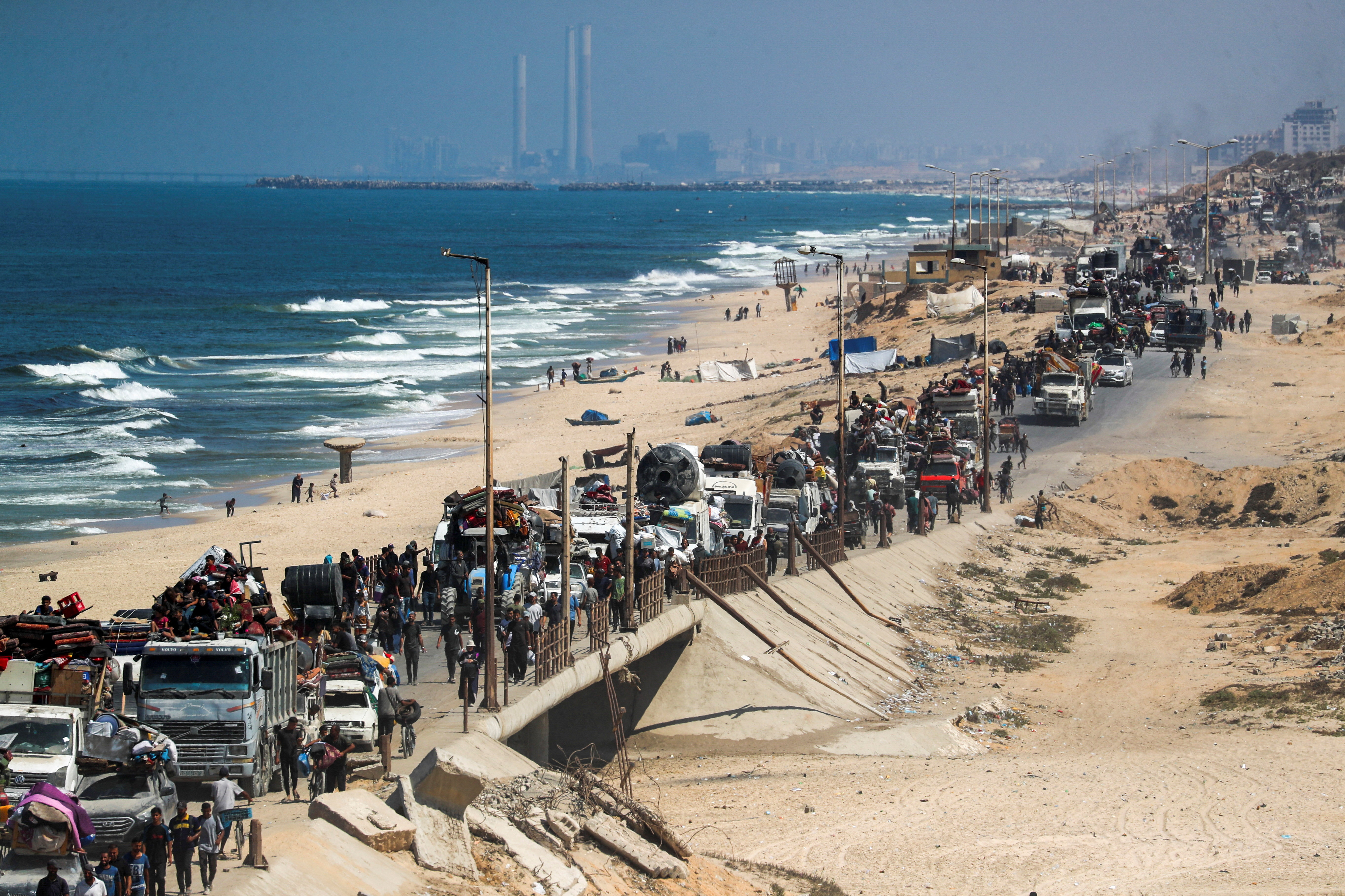 A long line of trucks loaded with people and belongings moves along a coastal road beside the sea, with more vehicles and crowds stretching into the distance.