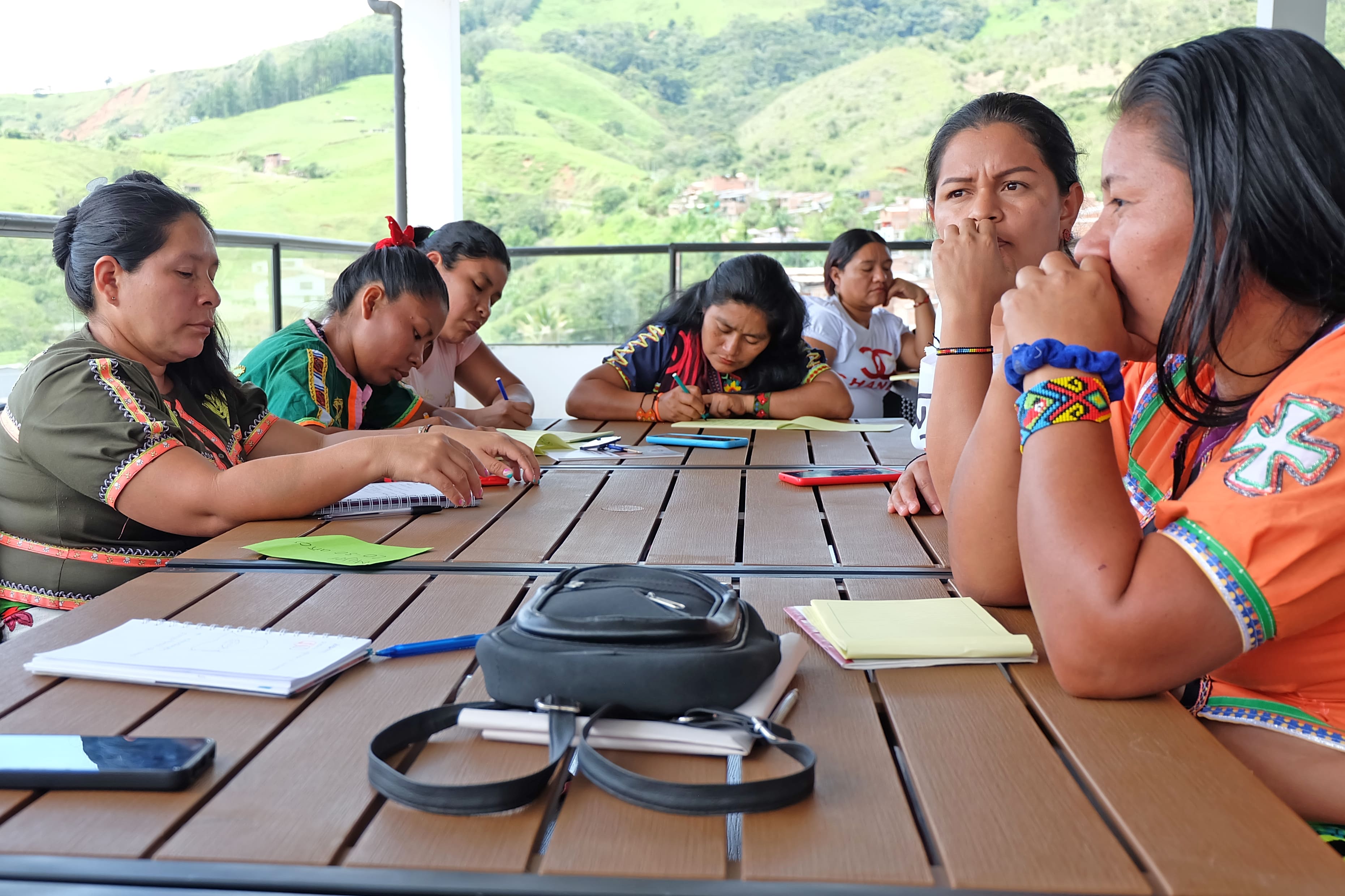 Un grupo de mujeres están sentadas escribiendo, y de fondo se ve un entorno rural