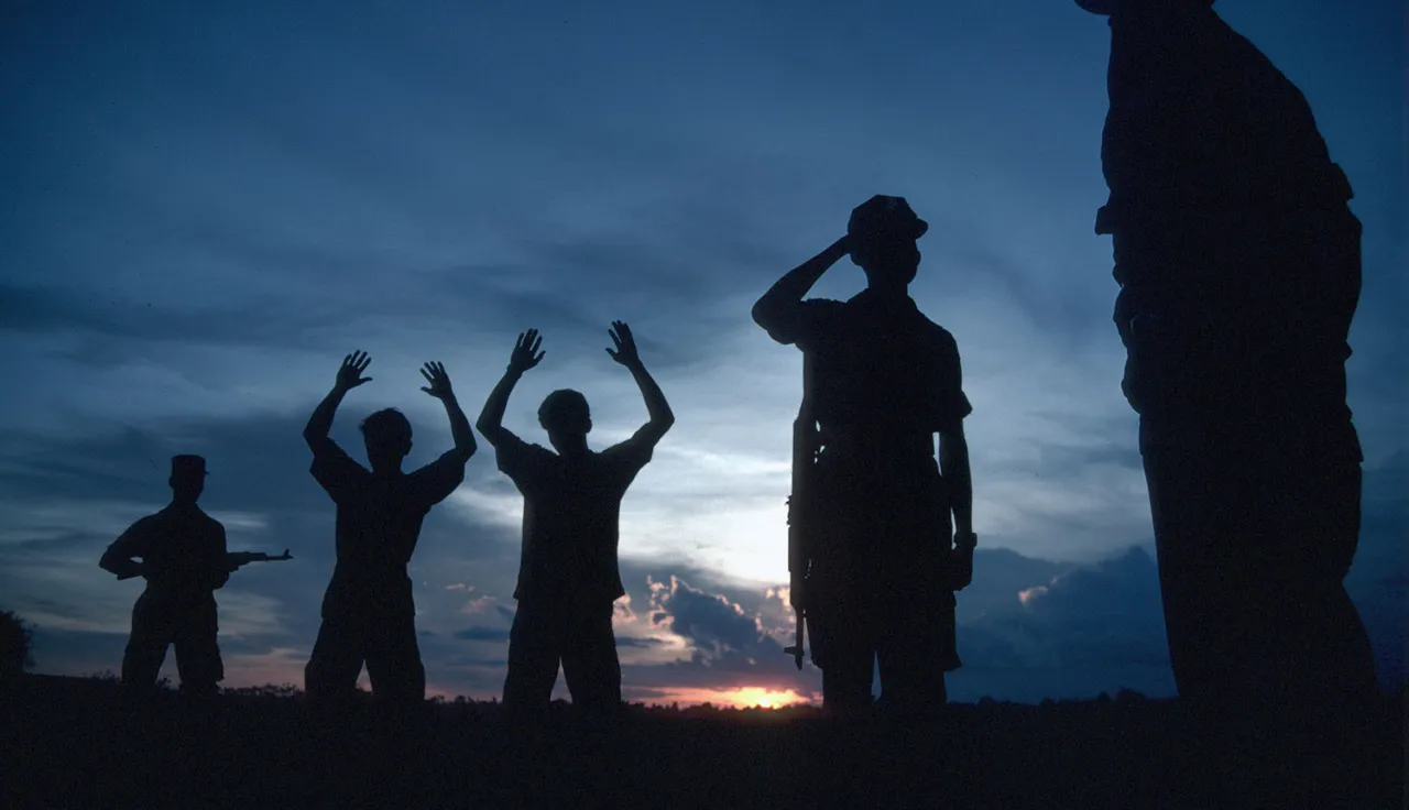 Two men standing with their hands raised up as another man points a weapon at them and two others look on