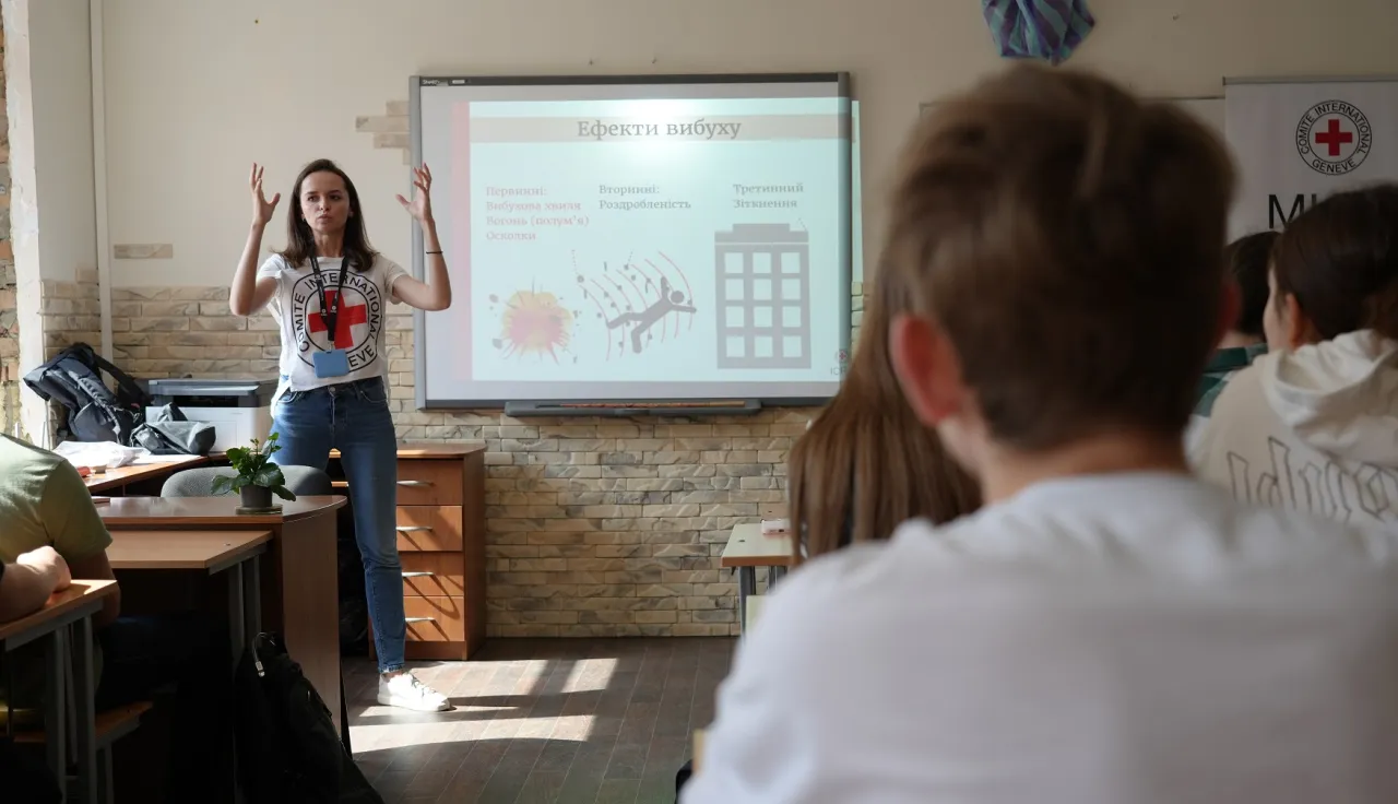 ICRC delelgate talking to students seated at desks in a classroom with information about explosive ordanances on a board behind her