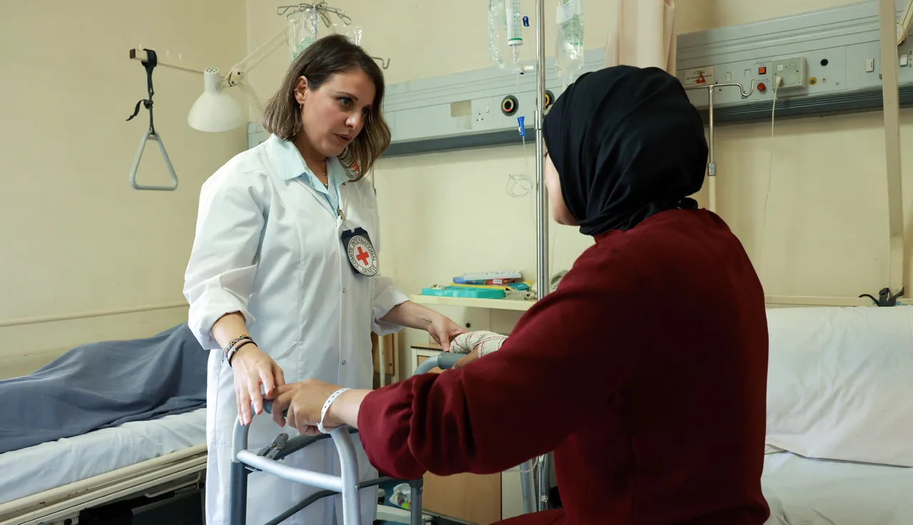 A woman sitting in a hospital bed holds a walker while talking to a female ICRC doctor 