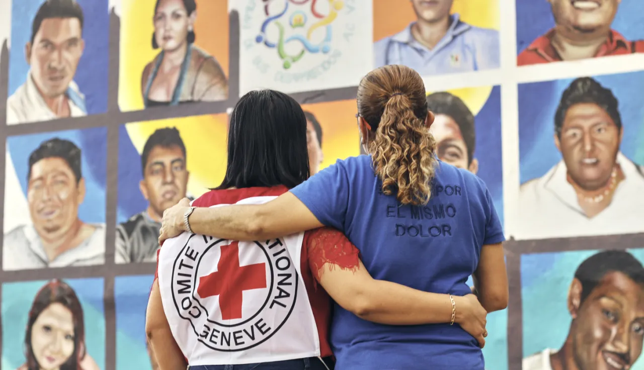 Una delegada del CICR y una familiar de una persona desaparecida se ven de espaldas abrazadas mirando un mural con ilustraciones de personas desaparecidas.