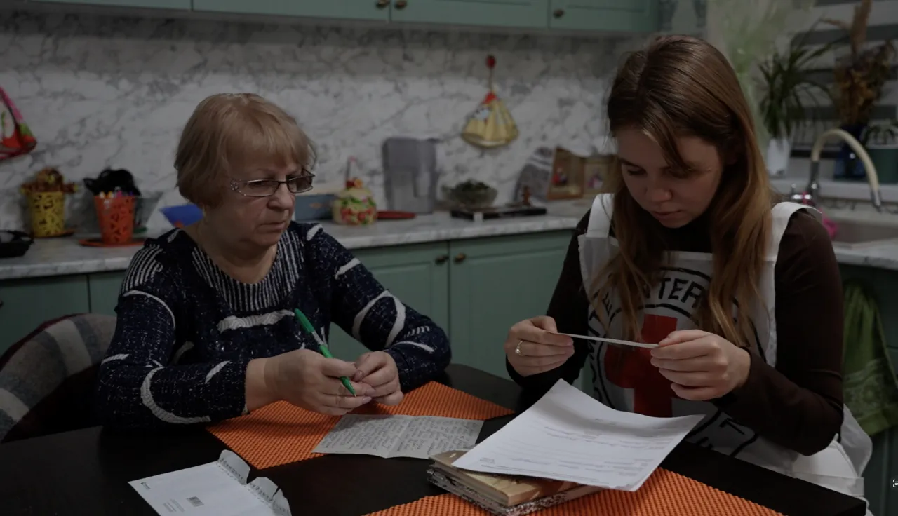 Madre de un prisionero de guerra sentada en un tanle con personal del CICR, escribiendo una carta a un ser querido.