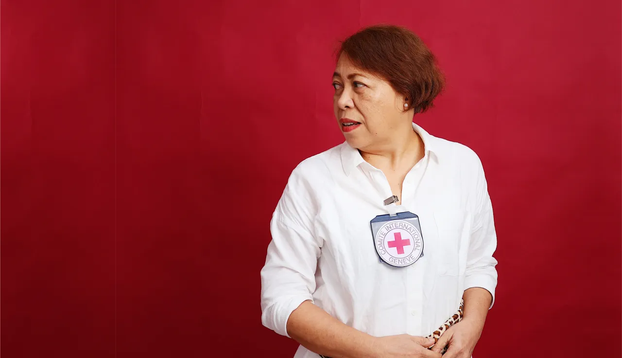 Mariegen Balo, in a white shirt with an ICRC badge, stands against a red background, looking to the side with a focused expression.