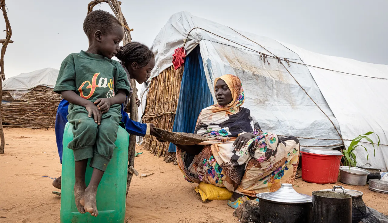 In a displacement camp in Sudan, a woman cooks over an open fire outside a makeshift shelter as two children watch.