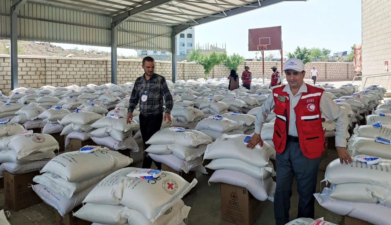 Two humanitarian workers (one from the ICRC and another one from the Yemeni Red Crescent) stand near each other surrounded by many piles of bags and boxes of food items