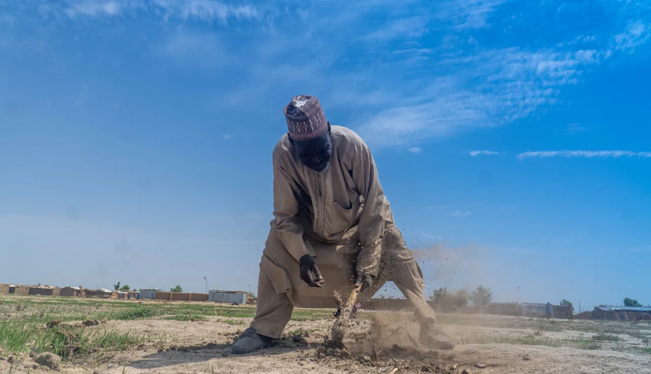 Churi Ibrahim, a 70-year-old farmer, works the dry, dusty soil with a hoe under a bright blue sky in a rural area, with modest shelters visible in the background — a scene reflecting resilience in the face of harsh living conditions.