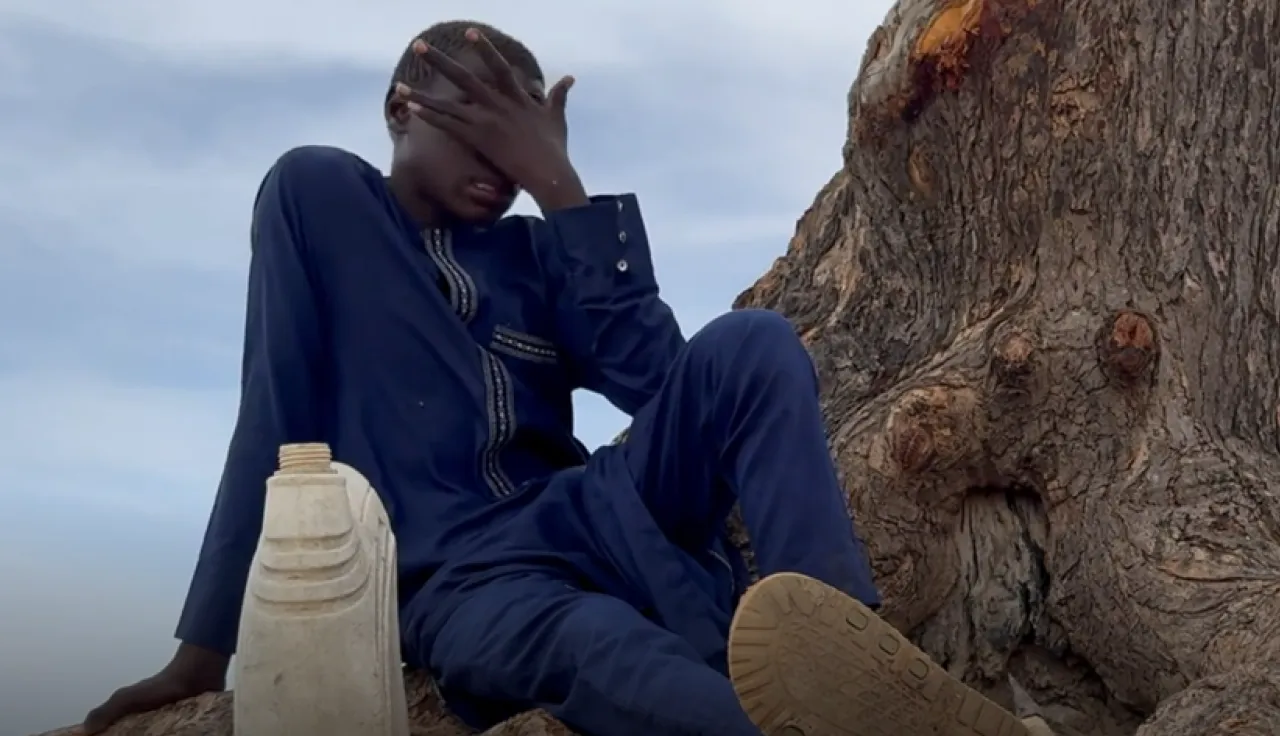 A young boy in a navy blue outfit sits at the base of a large tree, covering his face with one hand in a gesture of distress or exhaustion. A plastic water container rests beside him.