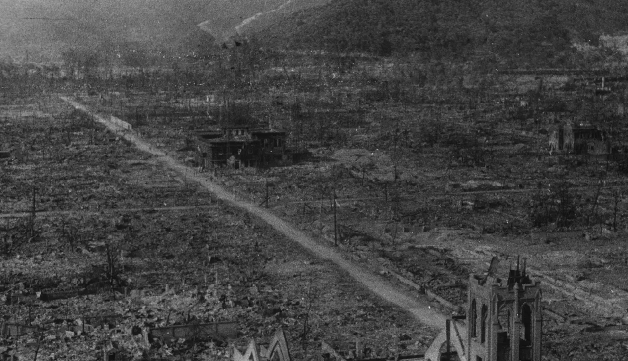 A black and white photograph showing the devastated landscape of Hiroshima after the nuclear bombing in August 1945. The area is flattened and burned, with only a few skeletal buildings and bare trees remaining. A straight dirt road runs through the center, surrounded by rubble and destruction, with mountains visible in the background.