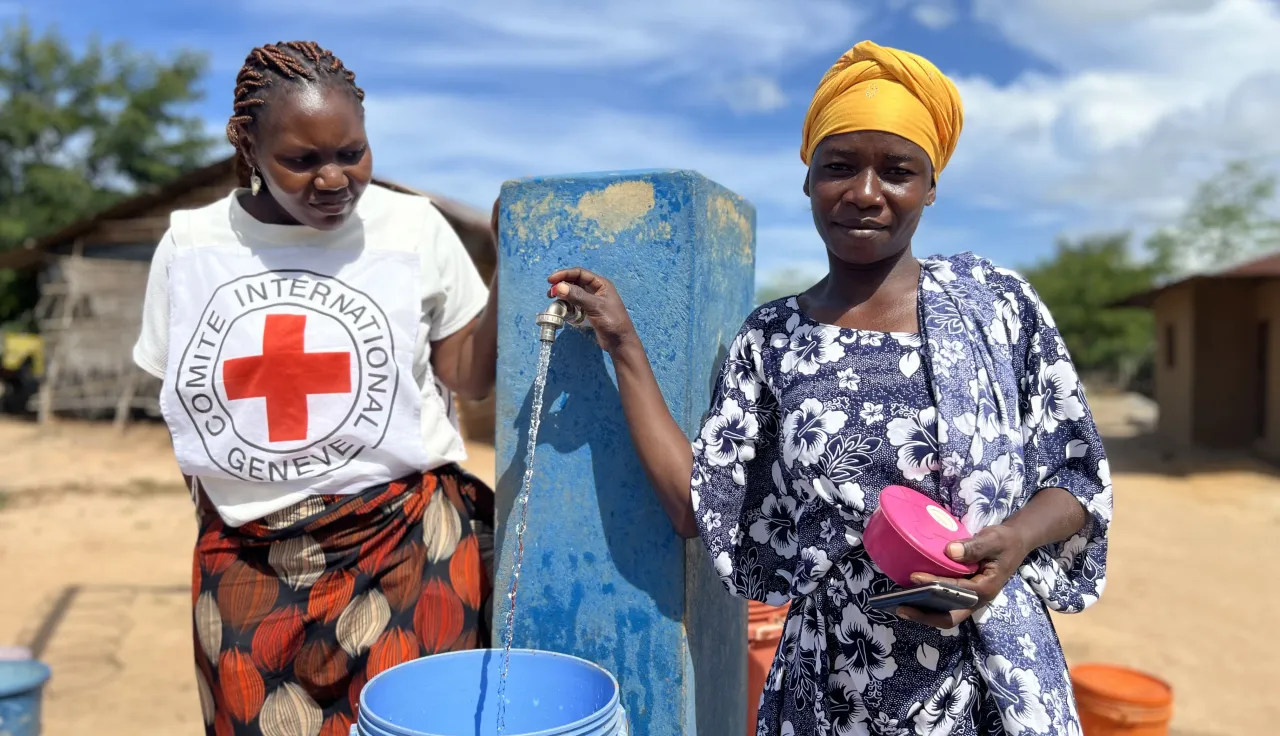 Mulher de uma comunidade enche recipiente de água numa fonte, ao lado de funcionária do CICV.