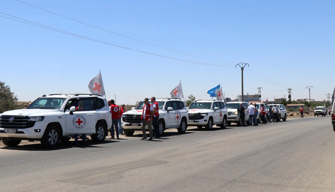 A convoy of white vehicles marked with Red Cross emblems and flags parked on a road under a clear blue sky, with humanitarian workers standing beside them.