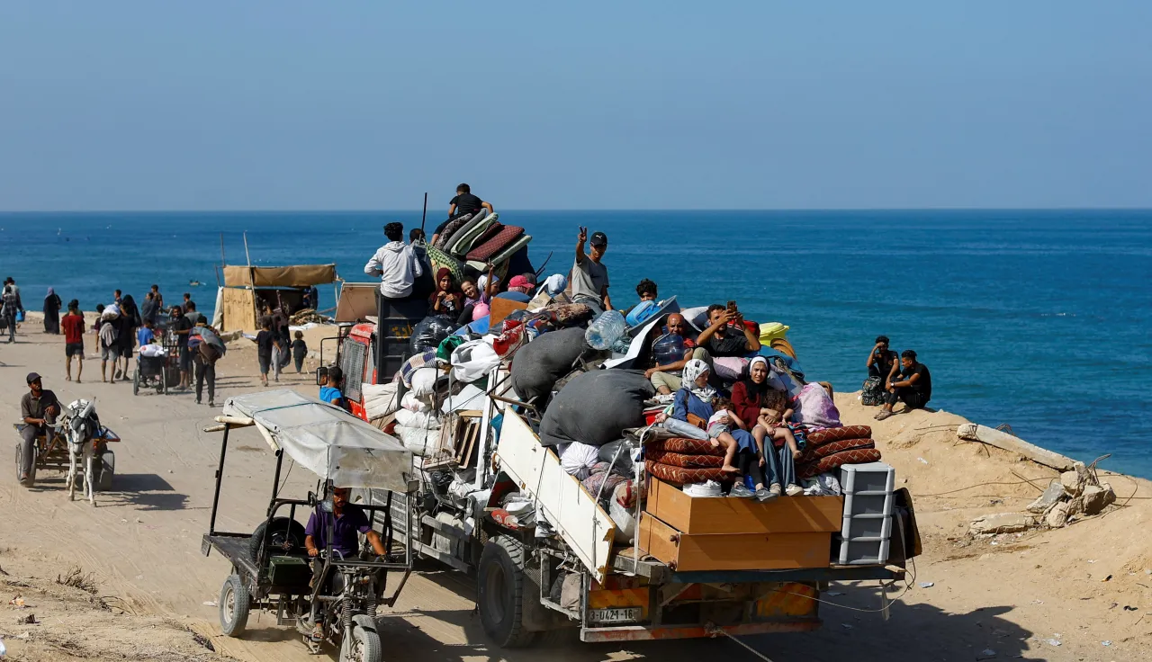 A crowded truck loaded with people and belongings drives along a coastal road, as others walk or travel on carts beside the sea.