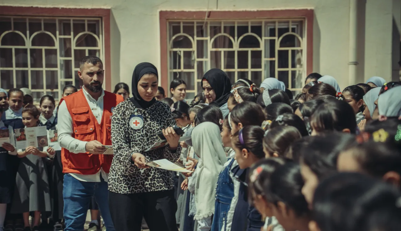 Two ICRC staff members interact with a group of primary school children. Some of the children are reading leaflets