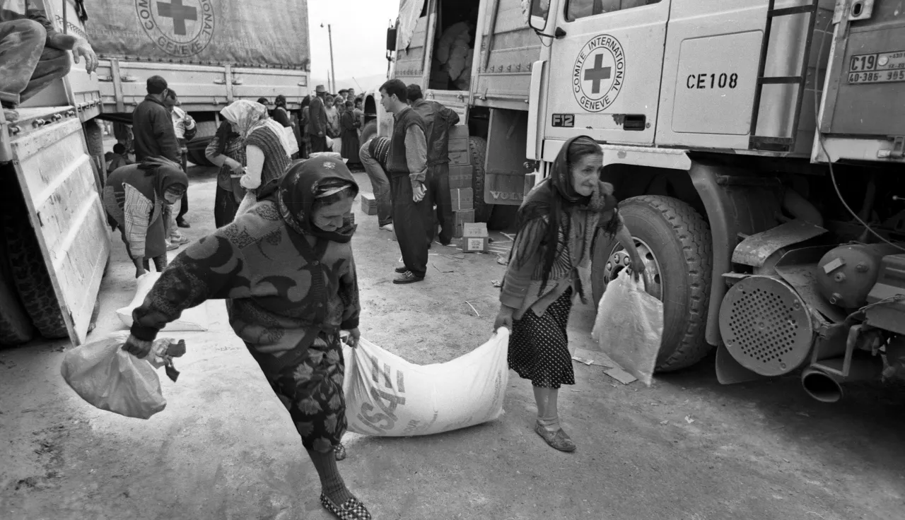 Two women carry a large sack of aid supplies together in front of Red Cross trucks, while other people unload and distribute relief items in the background