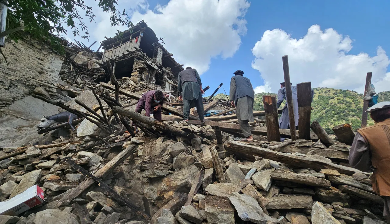 People search through the rubble of collapsed buildings after an earthquake, surrounded by broken wooden beams and stones, with mountains visible in the background under a partly cloudy sky.