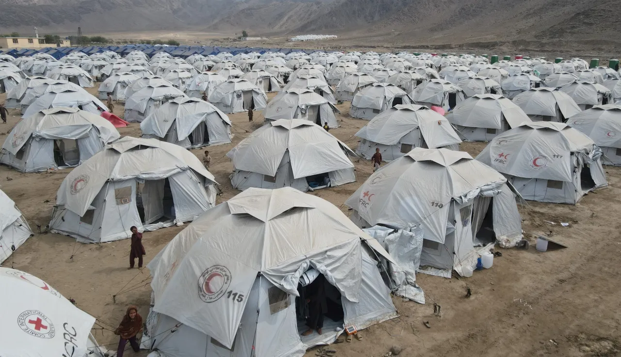 Rows of white humanitarian relief tents with Red Cross and Red Crescent logos set up in a camp on dusty ground, surrounded by mountains in the background.