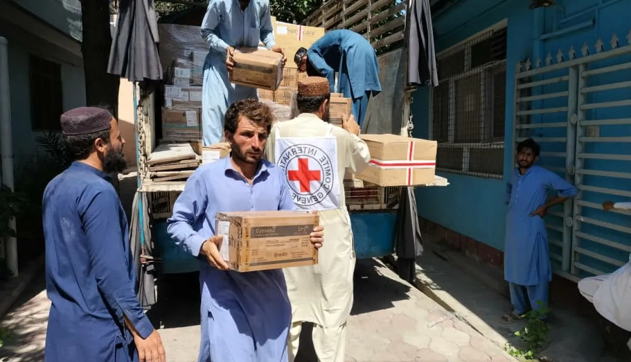 Humanitarian workers unload boxes of supplies from a truck, with one man wearing a vest marked with the Red Cross emblem, distributing aid outside a building.