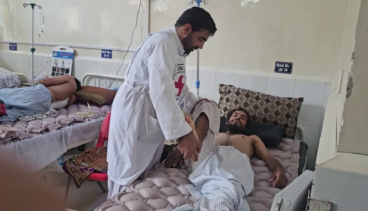 A medical worker in a white coat assists an injured man lying on a hospital bed, with other patients resting on beds in the background.