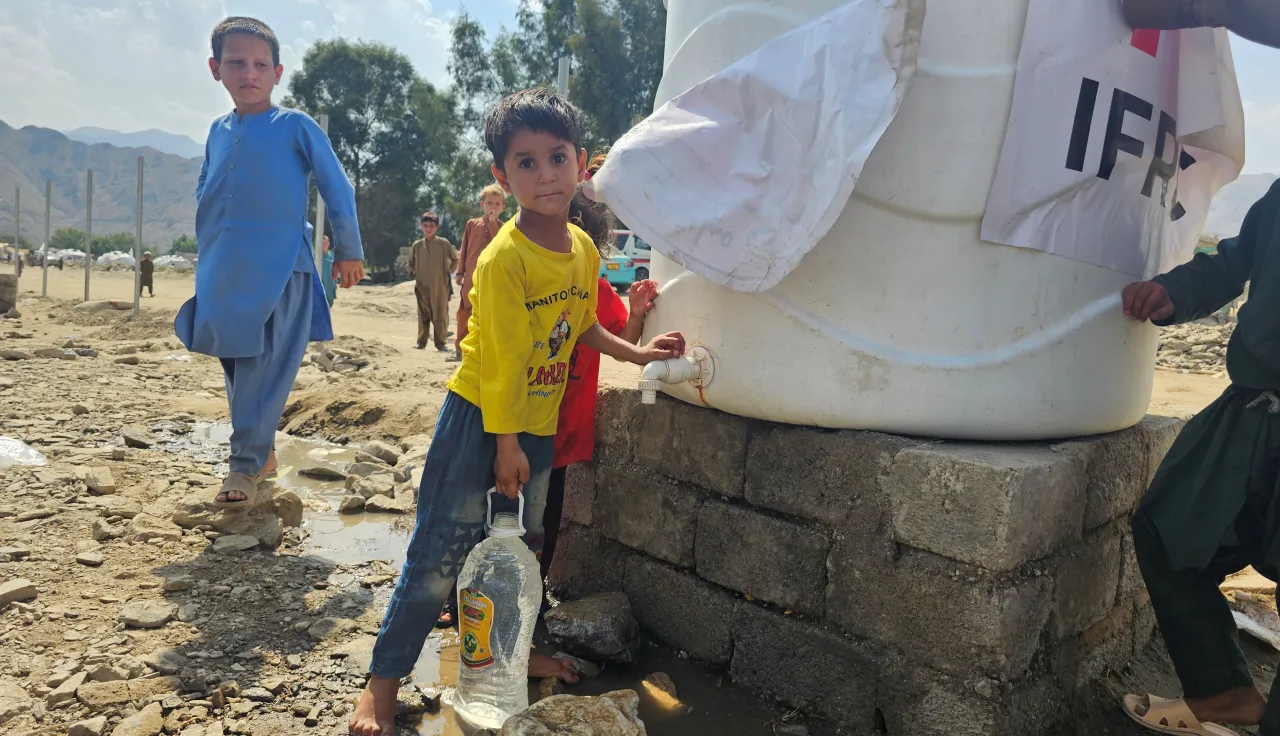 Children collect clean water from a large tank marked with humanitarian logos, one boy filling a plastic container, in a dry outdoor setting with mountains in the background.