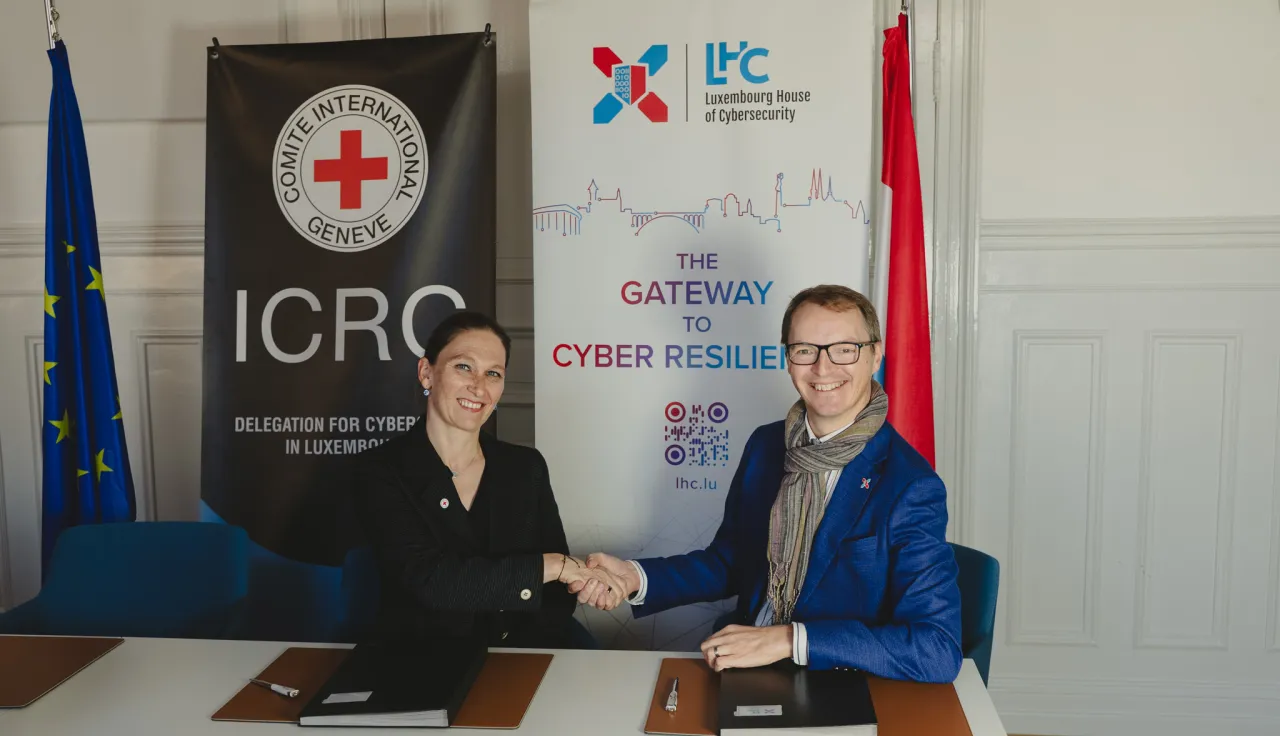 Head of ICRC Global Cyber Hub shaking hands with the CEO of Luxembourg House of Cybersecurity in front of the roll-ups of the respective organizations. With the flags of the European Union and Luxembourg next to the roll-ups.