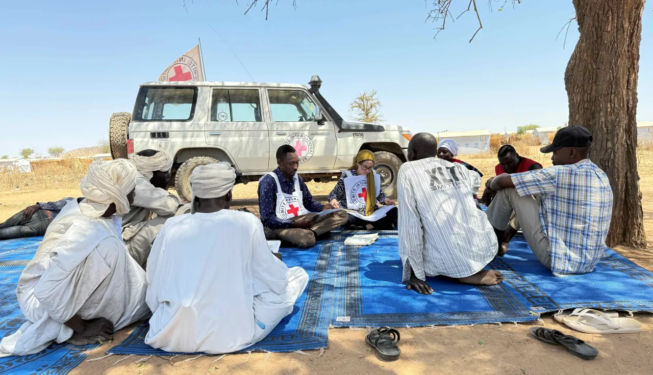 Des volontaires du Croissant-Rouge soudanais et des habitants de la communauté sont assis ensemble sur une bâche bleue, à l’ombre d’un arbre, discutant près d’un véhicule du CICR garé à proximité dans la région de Bara, au Soudan.
