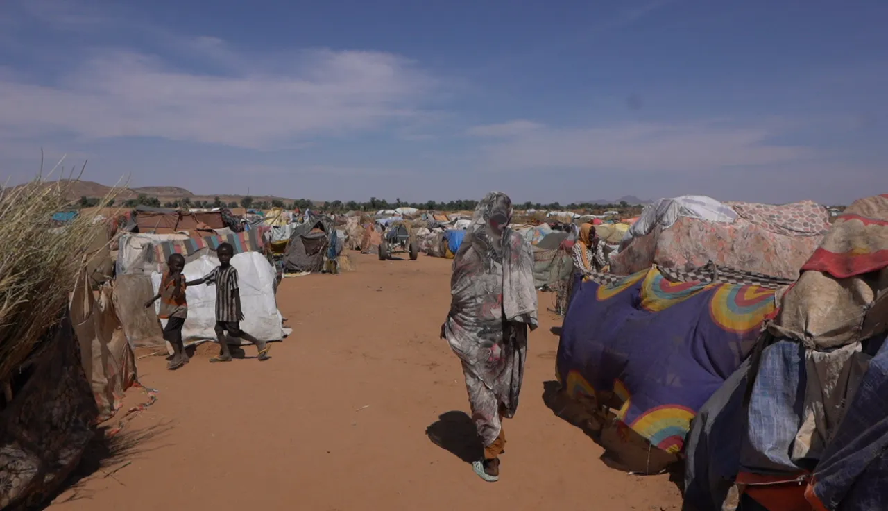 A wide view of a displacement camp in a dry, sandy area, with makeshift shelters covered in tarps and fabric stretching into the distance. Several people walk along the central pathway, including a woman in patterned clothing in the foreground and children on the left. The sky is clear and bright above the crowded camp.