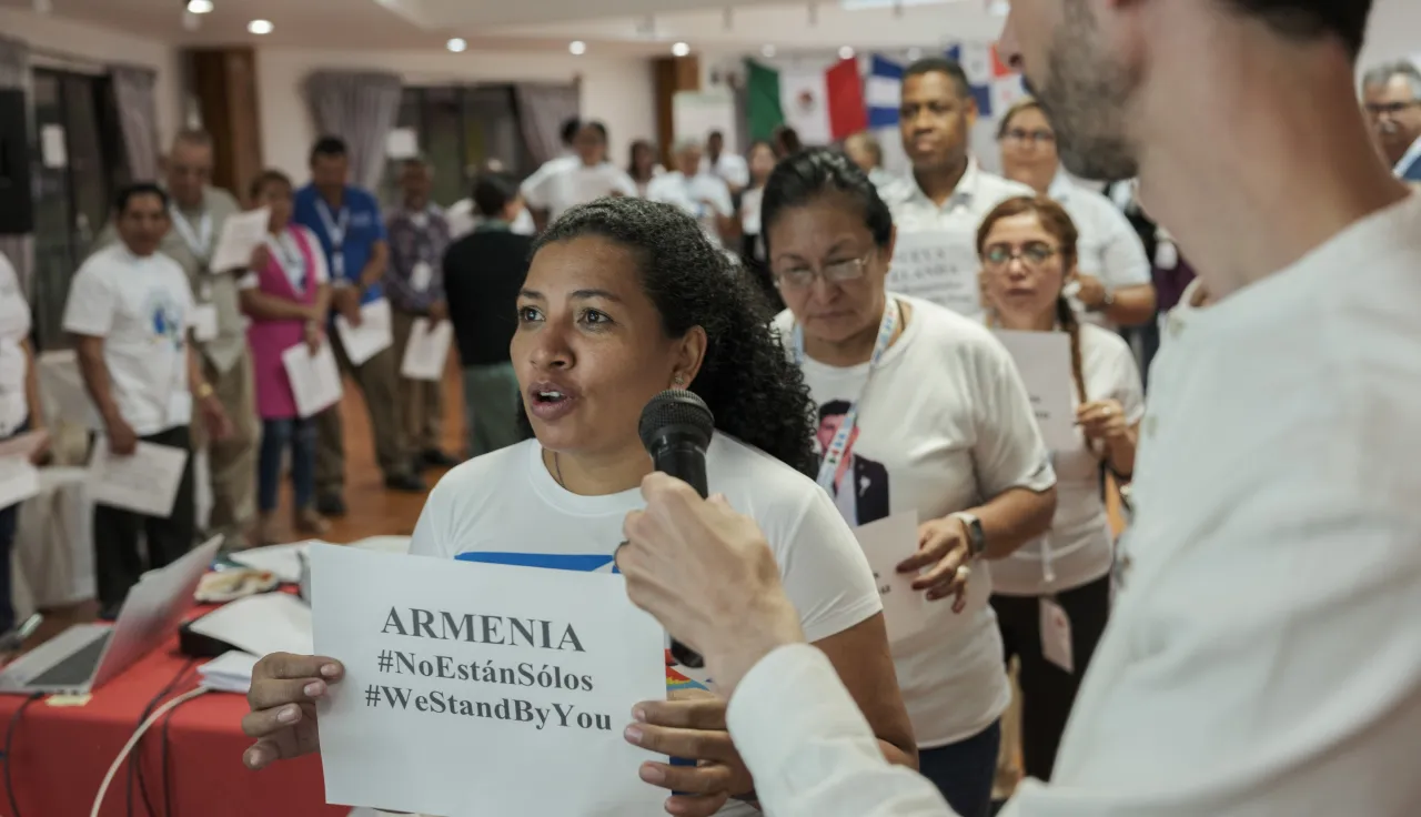A woman speaks into a microphone while holding a paper that reads “ARMENIA #NotAloneInSiles #WeStandWithYou,” during an indoor group event. Several people in white shirts stand behind her, some holding papers, with international flags visible in the background.