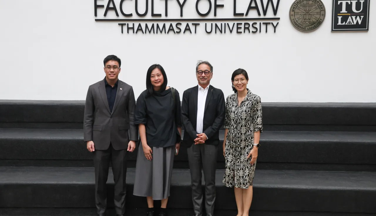 Four people standing and posing for a group photo in front of a wall that reads “Faculty of Law, Thammasat University,” with the university’s emblems displayed above them.
