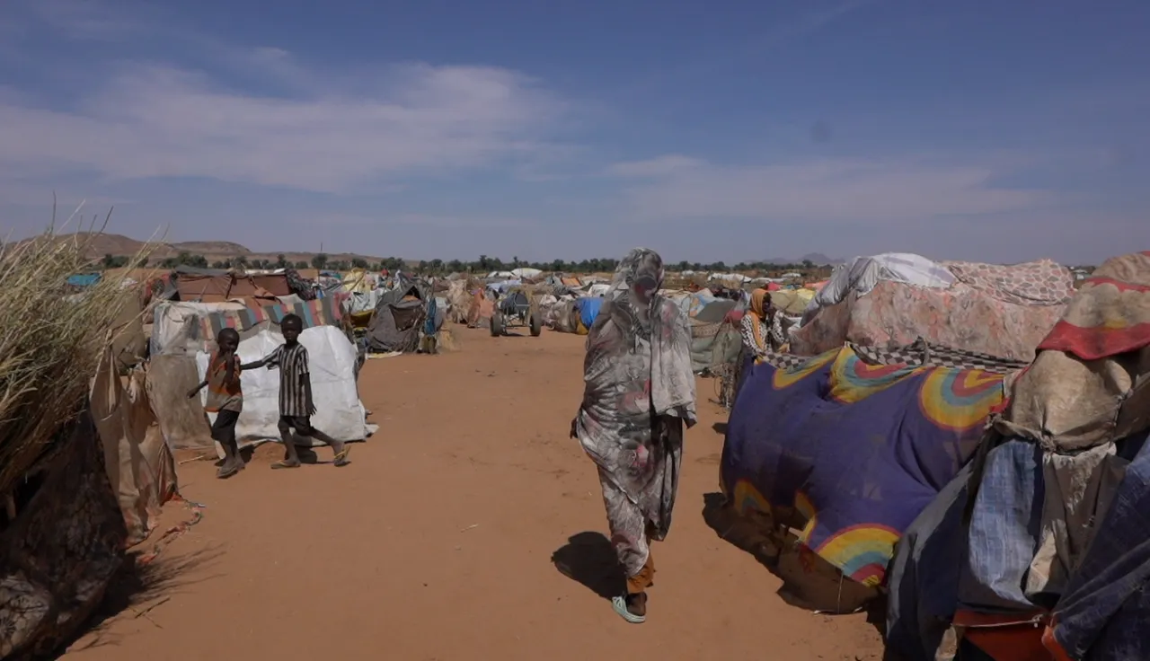 A woman walks through a dusty displacement site in Tawila, Sudan, passing makeshift shelters covered with tarps and fabric, while groups of people stand and move between the crowded structures under a clear blue sky.