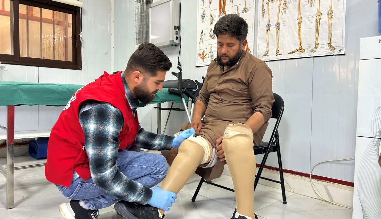 Joumaa, seated on a chair, wears two lower-limb prostheses as an ICRC staff member in a red vest kneels to adjust the fit. They are in a clinical rehabilitation room with anatomical posters on the wall.