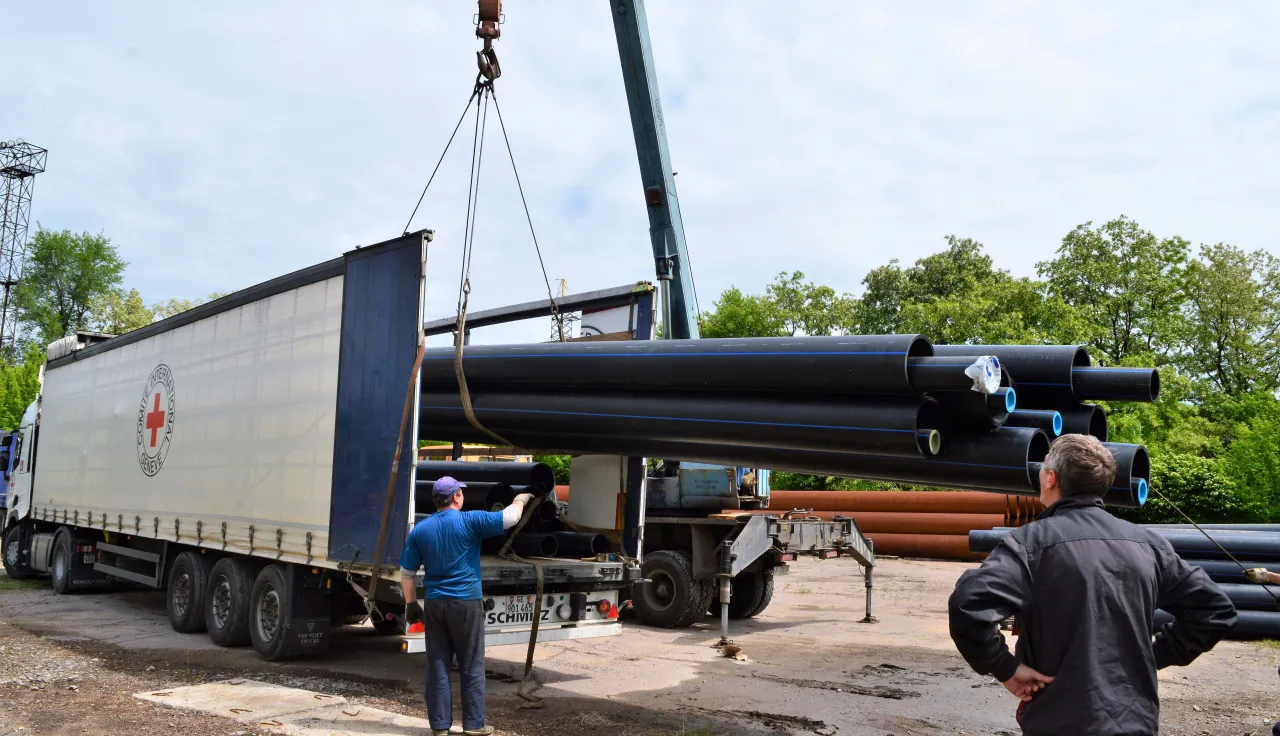 Workers use a crane to unload long black pipes from a truck at an outdoor work site, with one person guiding the load and others observing nearby.