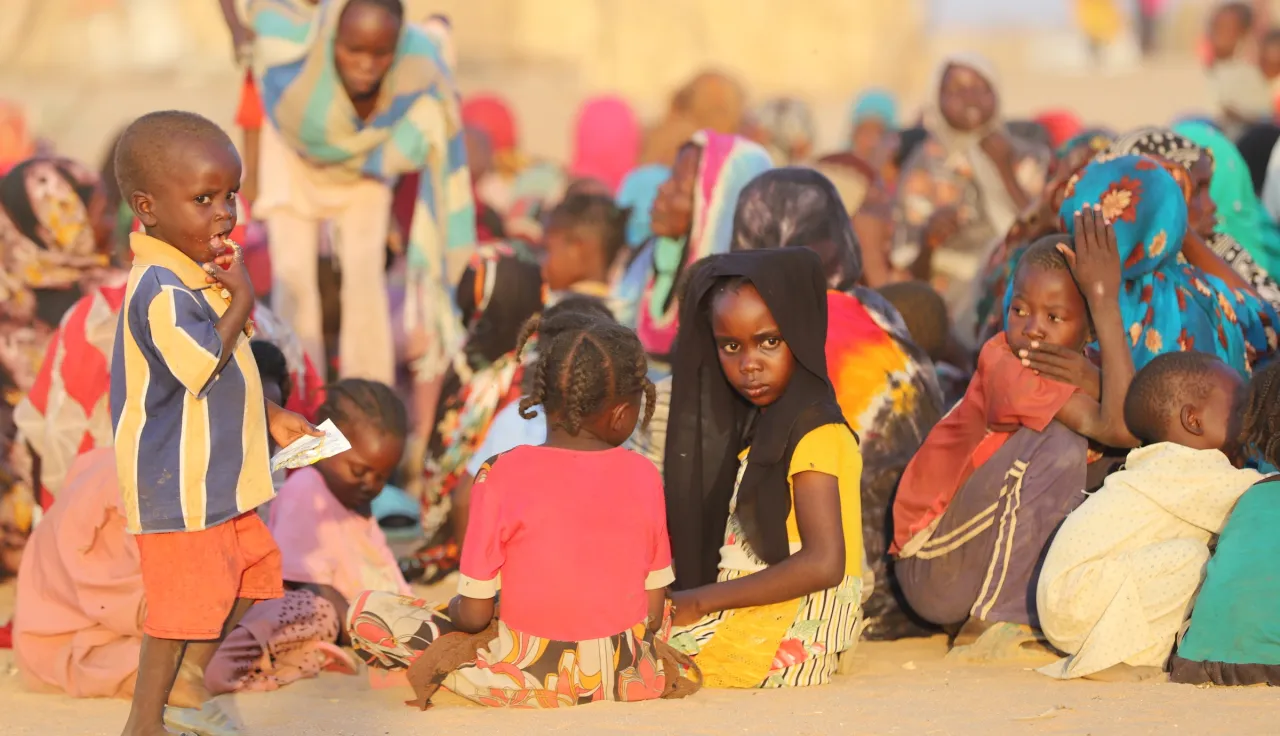 A group of displaced families sits together on sandy ground in Tawila, Sudan. Several women and children gather closely, some looking toward the camera. In the foreground, a young boy stands holding a piece of paper while another child in a headscarf sits among the group. The scene reflects the difficult conditions people are facing after fleeing violence.