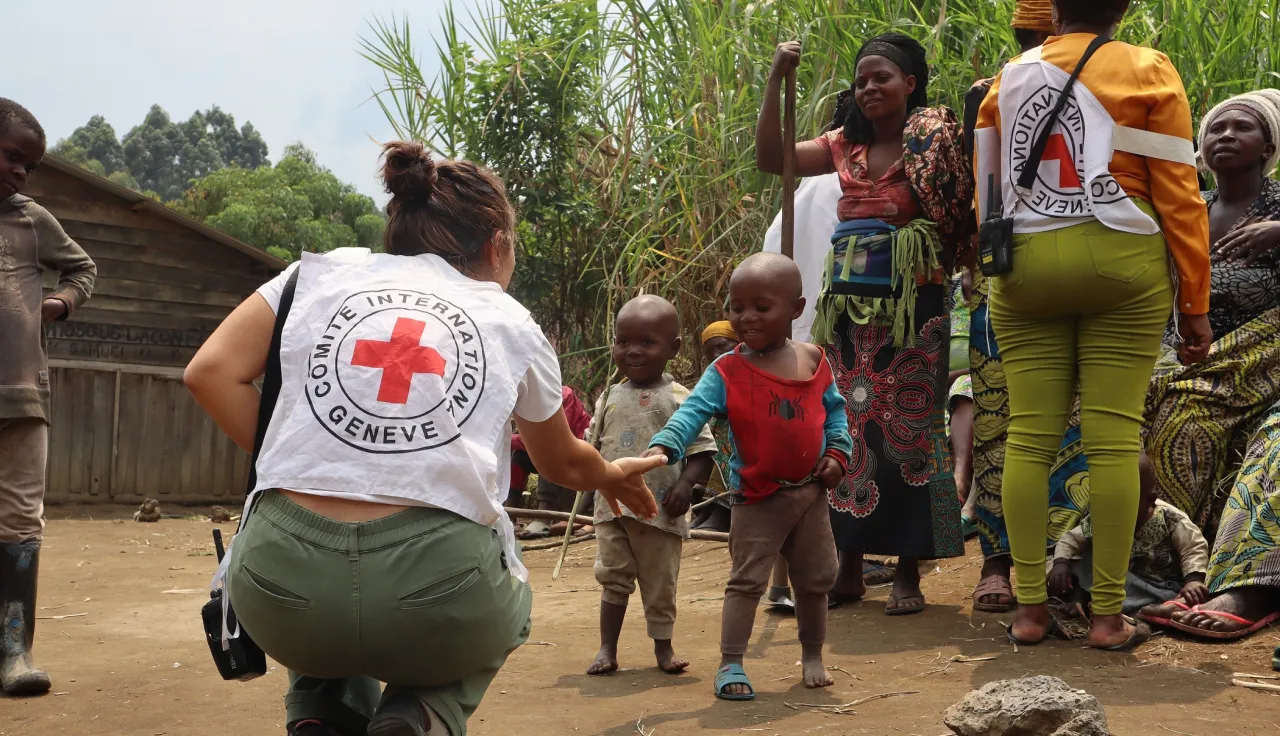 A woman reaches out her hand to two children, with other women standing in the background.