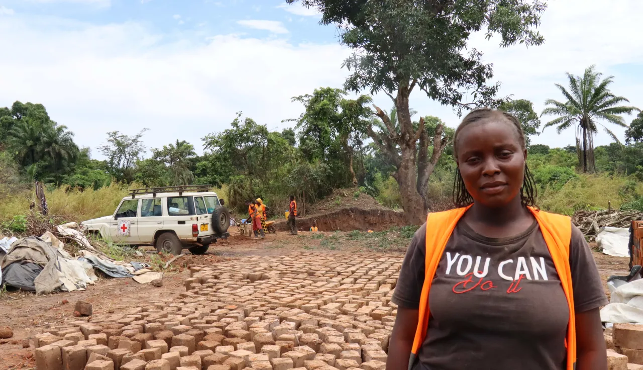 Una mujer mira a la cámara. A sus espaldas hay ladrillos, más personas de la comunidad y un carrod del CICR.