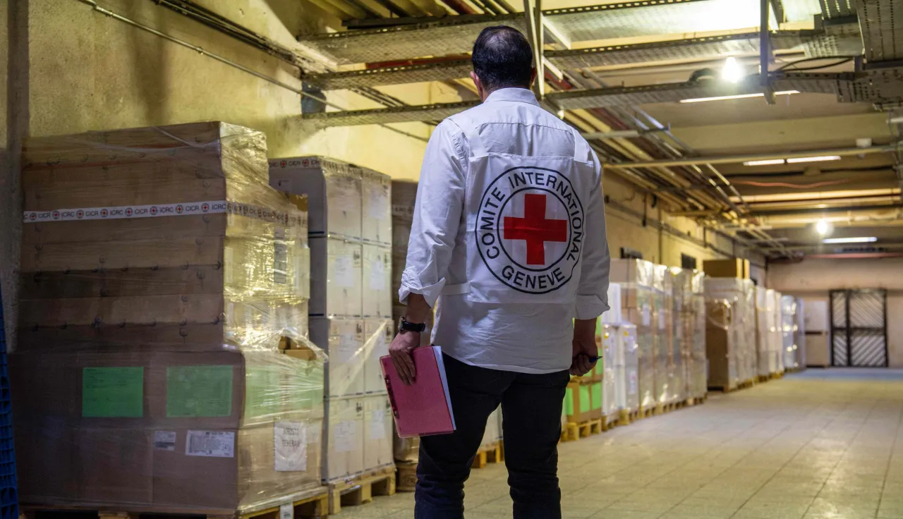 ICRC staff member wearing a white vest with the red cross emblem walks through a warehouse lined with pallets of boxed humanitarian supplies, carrying documents in hand.
