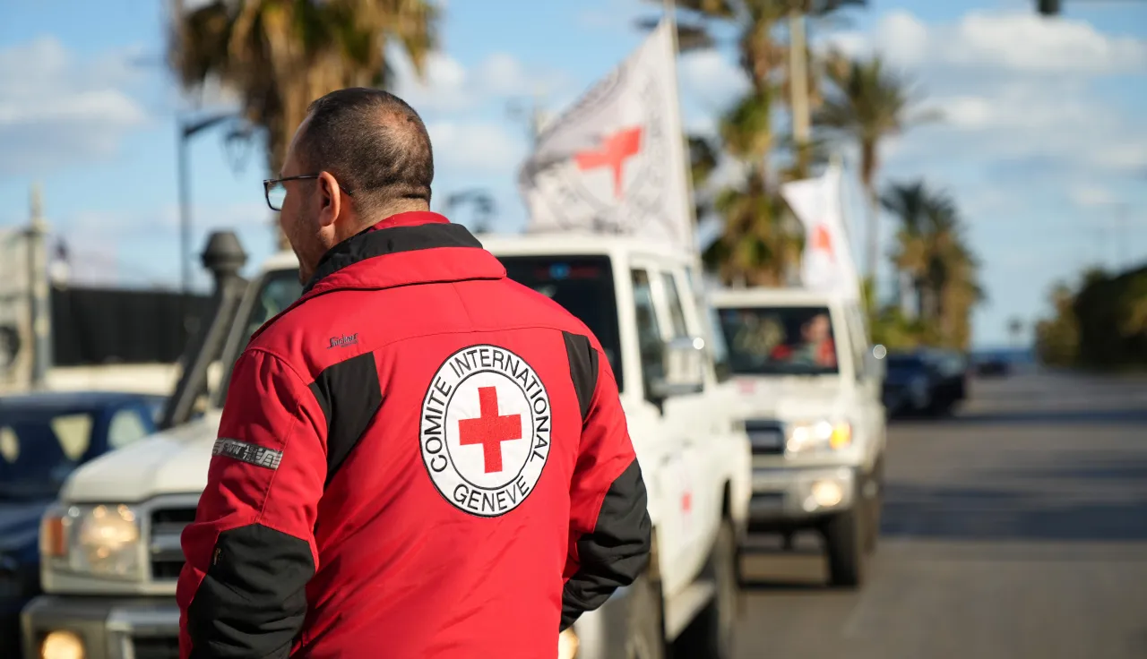 An ICRC staff member wearing a red jacket with the Red Cross emblem stands beside a line of Red Cross vehicles on a coastal road in Lebanon during humanitarian operations amid escalating hostilities.