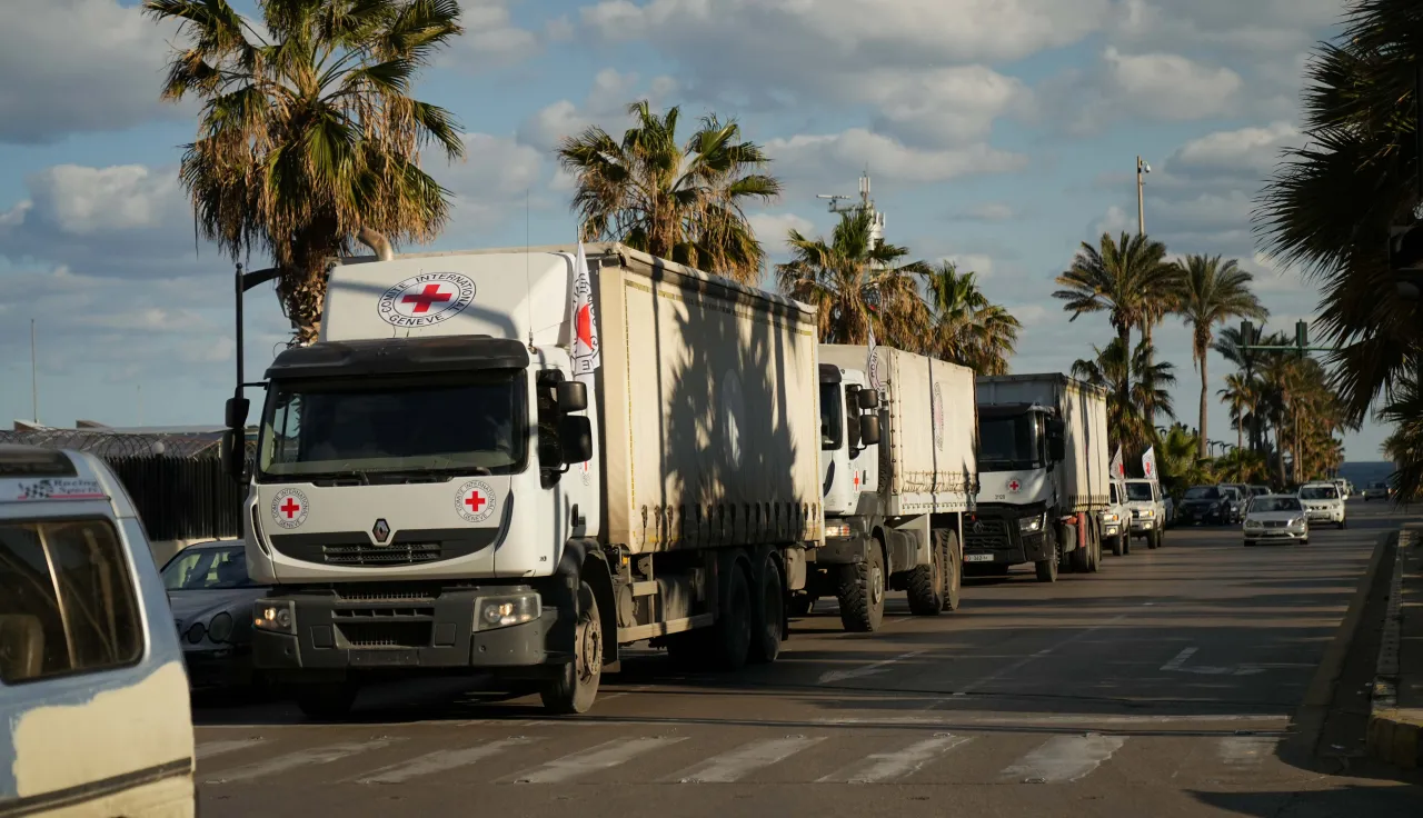 A convoy of trucks bearing the Red Cross emblem drives along a palm-lined coastal road in Lebanon, delivering humanitarian supplies during escalating hostilities.