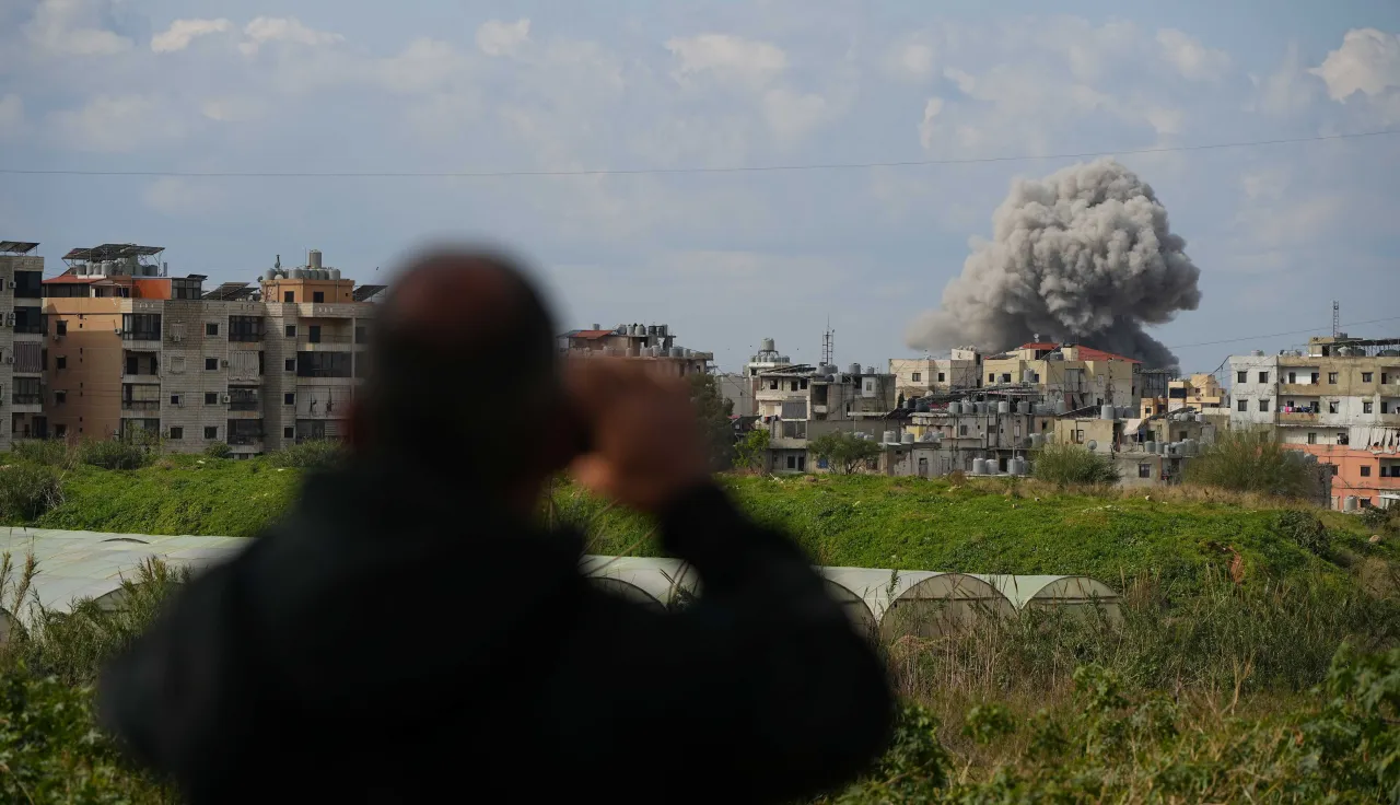 A person stands in the foreground, blurred and seen from behind, holding a phone while looking toward a city where a large plume of smoke rises above buildings in the distance.