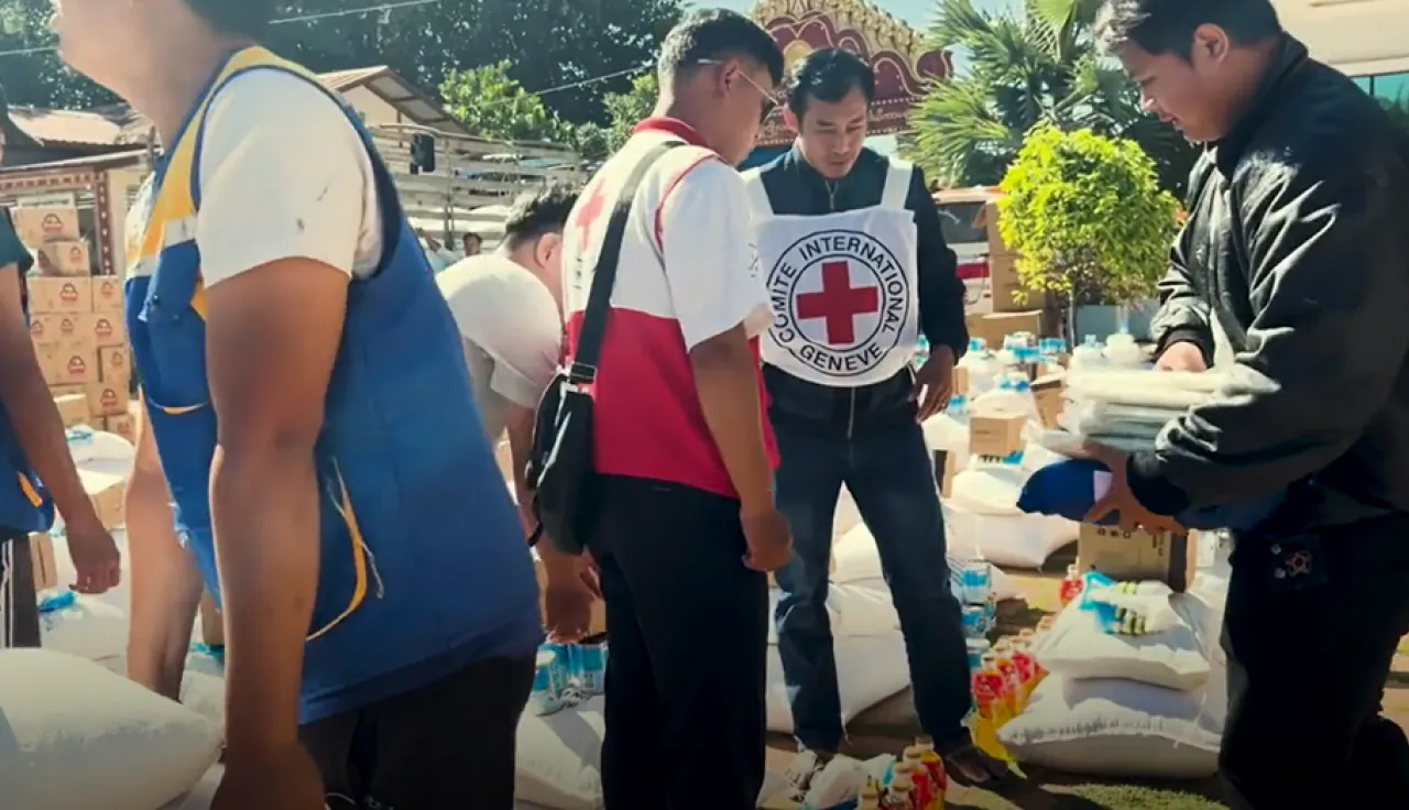 Aid workers distribute relief supplies to people gathered outdoors, with a Red Cross volunteer assisting at a table stacked with bags of goods.