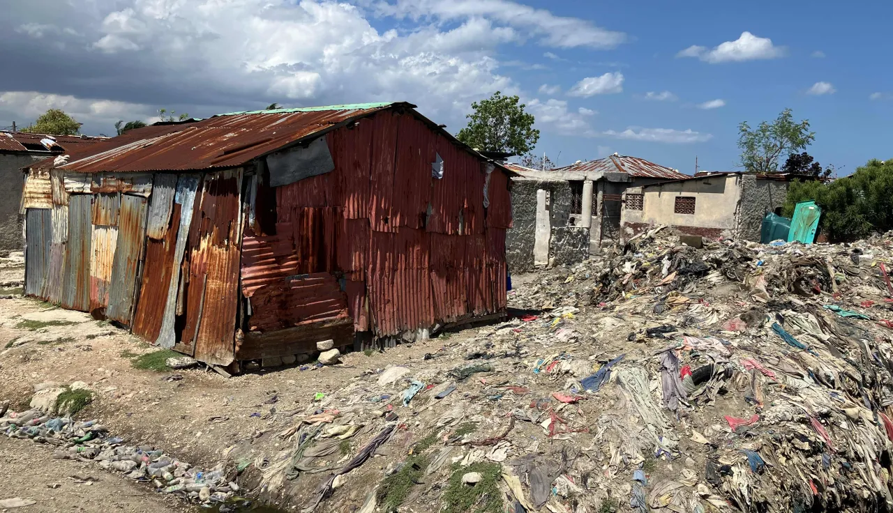 Vivienda construida con láminas de metal oxidadas se encuentra junto a un terreno cubierto de basura y escombros. Alrededor hay otras estructuras, bajo un cielo azul con nubes dispersas.