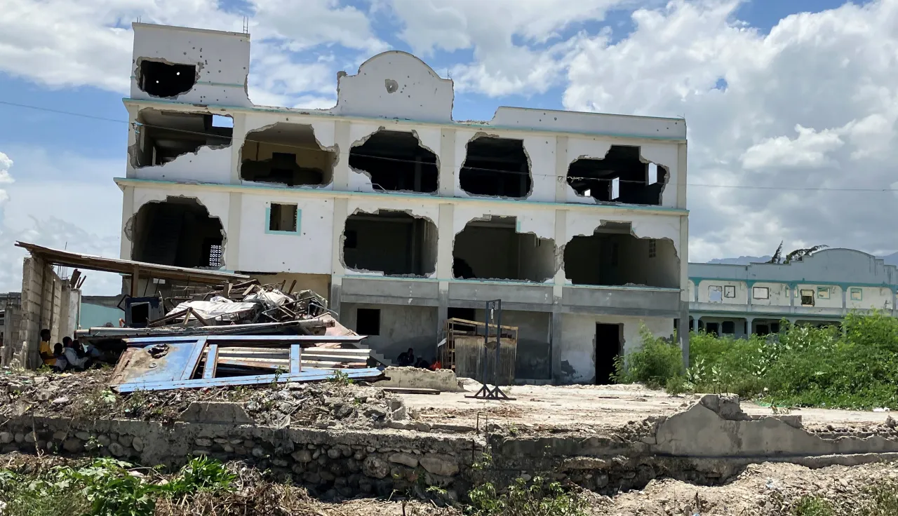 Edificio de varios pisos parcialmente destruido, con ventanas y paredes abiertas, se encuentra en un entorno con escombros y estructuras colapsadas en primer plano. El área circundante muestra vegetación dispersa y otros edificios al fondo bajo un cielo con nubes.
