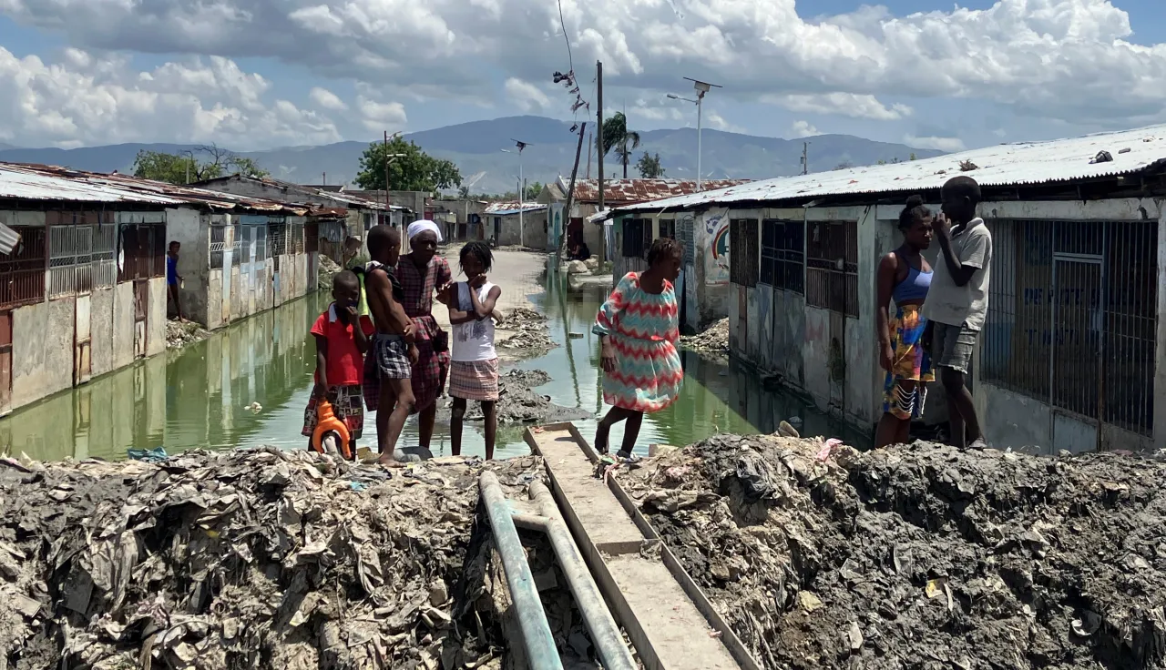 Personas, entre ellas varios niños, se encuentran en un asentamiento con viviendas precarias, donde el agua estancada ha inundado el área. Un estrecho paso improvisado con tablas cruza sobre el agua, mientras las casas de ambos lados muestran condiciones deterioradas bajo un cielo parcialmente nublado.