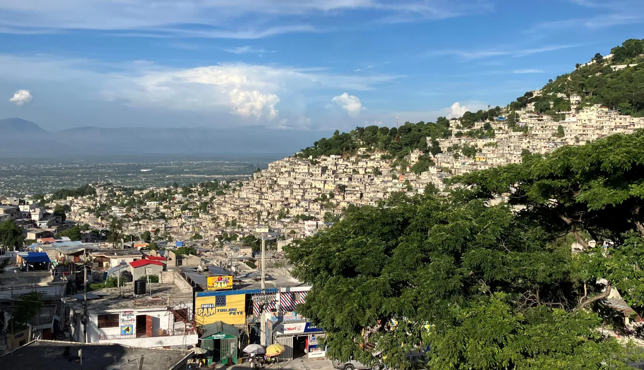 Vista panorámica de Puerto Príncipe, Haití, donde viviendas densamente agrupadas de colores claros se extienden por una ladera montañosa. En primer plano se observan techos y árboles, mientras al fondo se aprecia el valle bajo un cielo parcialmente nublado.