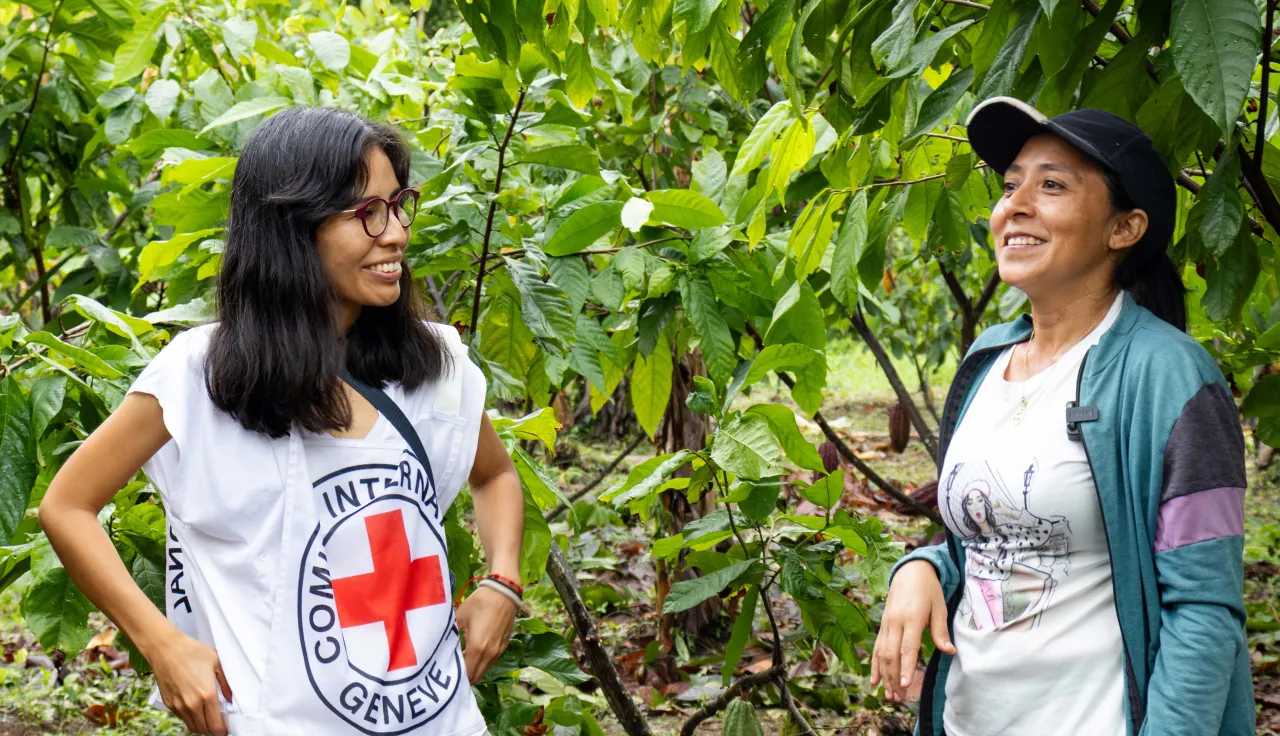 Dos personas conversan al aire libre en un entorno verde con plantas. Una lleva chaleco blanco con el emblema del Comité Internacional de la Cruz Roja. La escena ocurre en Apure, Venezuela, durante una visita a proyectos de medios de vida.