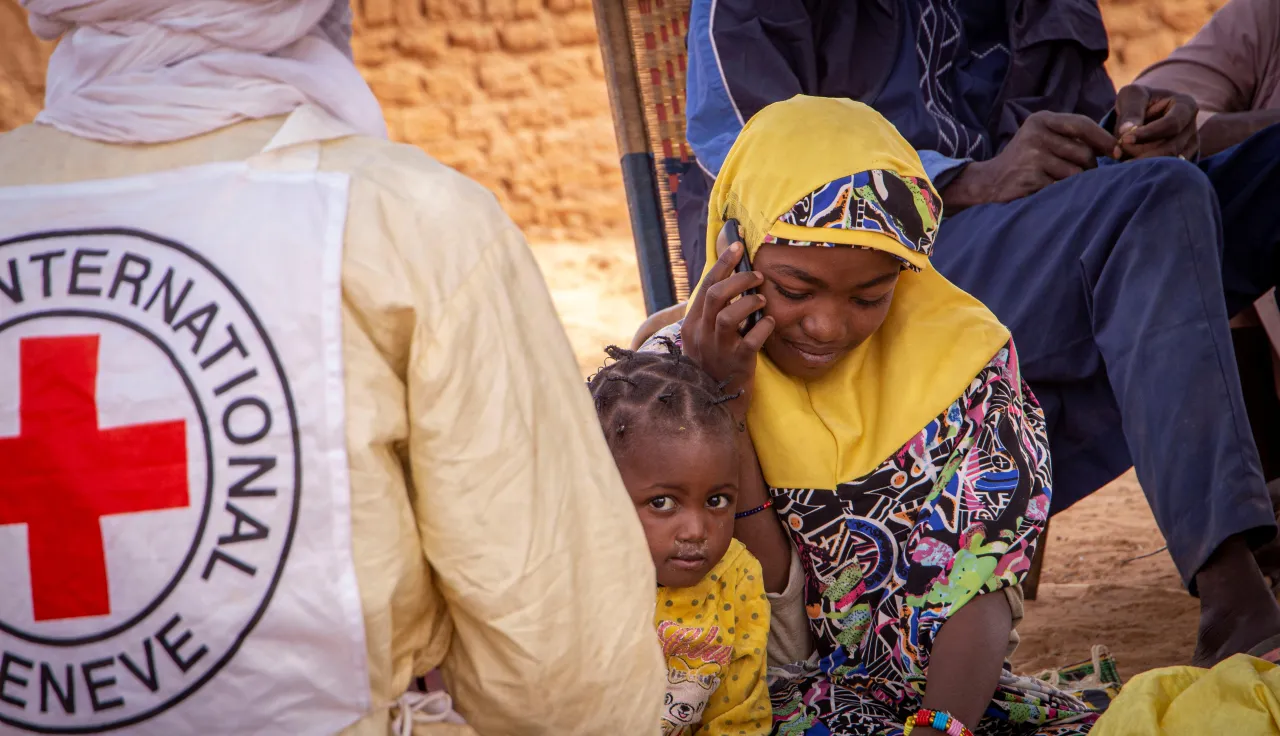 A woman wearing a yellow headscarf sits and speaks on a phone, with a young child beside an ICRC vest in Anderamboukane, Mali, as people gather in the background outdoors.