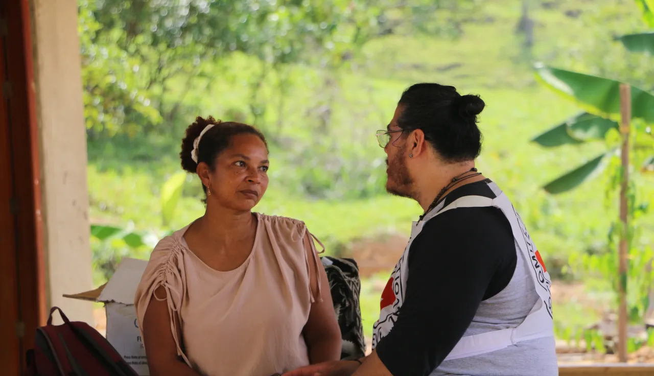 Una persona del Comité Internacional de la Cruz Roja conversa con una persona de la comunidad en una zona rural. Ambos están de pie al aire libre, rodeados de vegetación y paisaje campestre.