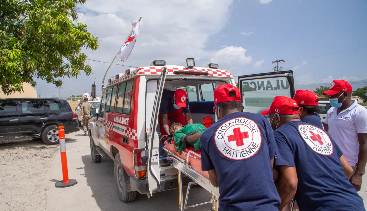 Personal de la Cruz Roja Haitiana traslada a una persona herida en camilla hacia una ambulancia. Varios socorristas participan en la evacuación y atención de emergencia durante el traslado del paciente.