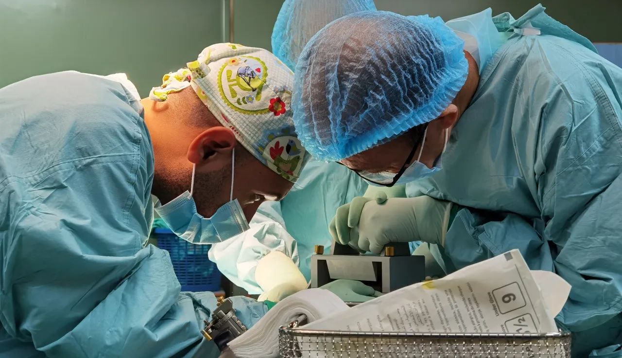 Two surgeons in blue protective gowns and caps perform a delicate operation under bright surgical lights, focusing intently over a patient on the operating table.