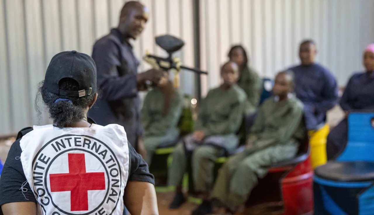 An ICRC staff member oversees a practical session of Law Students during an armed conflict simulation excersise on IHL. She is looking at an instructor holding a mock rifle as students are seated looking attentively looking at the instructor.